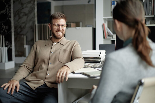 Caucasian young adult man smiling and talking with Caucasian young adult woman in modern office setting, both sitting at desk with stack of books and open notebook visible