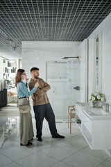 Caucasian young woman and man standing in modern bathroom showroom taking photo on smartphone