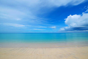 Calm turquoise ocean water meets a sandy beach under a vast blue sky with scattered white clouds, creating a serene natural landscape.