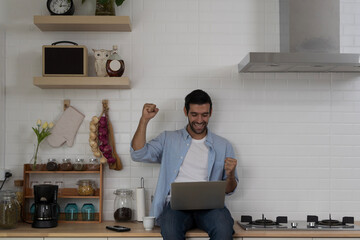 Happy young man celebrating success while using laptop in modern kitchen, joyful remote work...