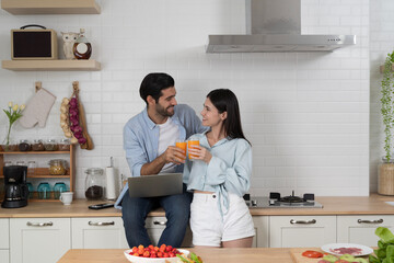 Smiling couple drinking fresh orange juice together in modern kitchen with laptop, love lifestyle concept