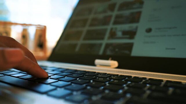 Close-up of a person typing on a laptop keyboard with a blurred screen showing images
