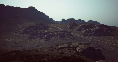 Hikers traverse a winding path through rocky hills as dusk approaches. Shadows stretch across the landscape, creating a dramatic atmosphere and highlighting the natural beauty.