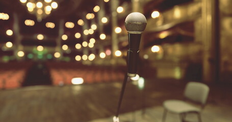 A microphone stands in focus at a beautiful theater stage, with soft lights illuminating the background. A lone chair sits nearby, suggesting a performance is about to begin.