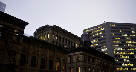 Buildings in an urban area are illuminated as dusk approaches, showcasing a mix of old and modern architecture. A water tower stands atop one of the structures.