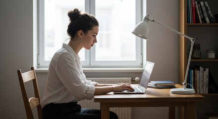 Focused on Work: A young woman, immersed in concentration, sits at her wooden desk, her fingers dancing across the keyboard as she works on her laptop by a sunlit window.