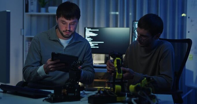 Team adult male engineer collaborate on robotic hand project using AI code solutions machine learning at tech workspace night. Artificial intelligence interface, collaboration prototype development.