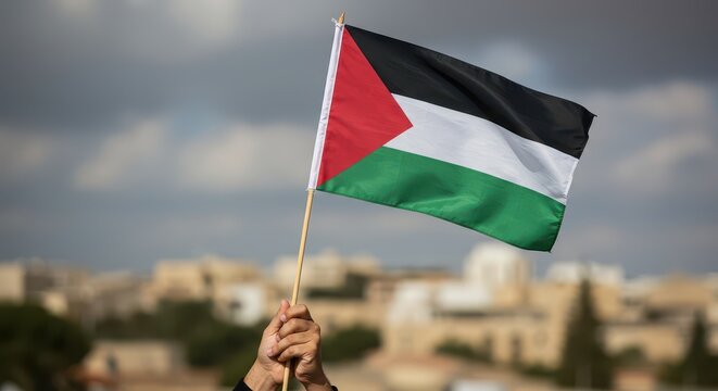 Waving Palestinian Flag: A hand triumphantly waves the flag of Palestine, symbolizing national pride, heritage, and identity against a blurred architectural backdrop.