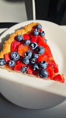 Slice of berry fruit tart with blueberries and strawberries on a white plate in natural light