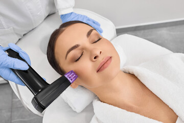 Brunette woman receiving radiofrequency lifting procedure for her face skin rejuvenation at...