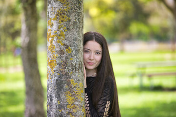 Latin woman, young and beautiful brunette dressed in black is clinging and hiding behind a tree...