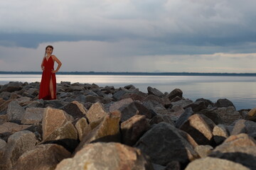 Pretty young brunette woman in red dress walking by rocks at the seashore