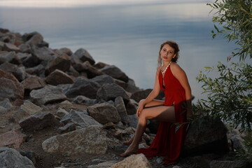 Slender long legged barefoot beautiful brunette young woman in long red dress sitting on the rocks of the sea coastline