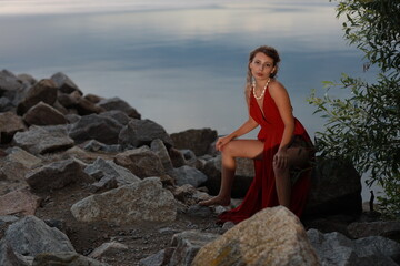 Slender long legged barefoot beautiful brunette young woman in long red dress sitting on the rocks of the sea coastline