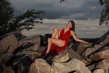 Slender long legged barefoot beautiful brunette young woman in long red dress sitting on the rocks of the sea coastline