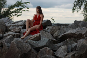 Slender long legged barefoot beautiful brunette young woman in long red dress sitting on the rocks of the sea coastline