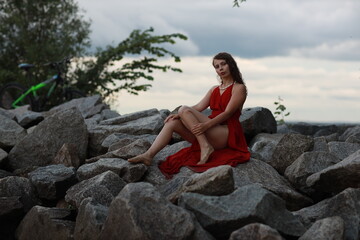 Slender long legged barefoot beautiful brunette young woman in long red dress sitting on the rocks of the sea coastline