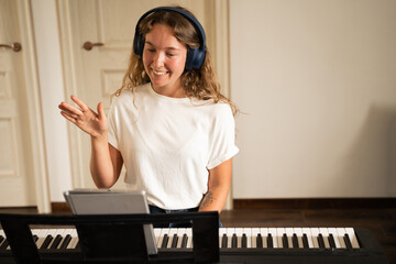 Front view of happy young woman taking online piano lessons at home using tablet waving her hand,...