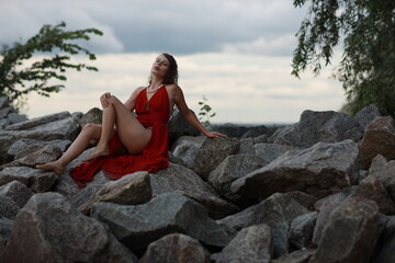 Slender long legged barefoot beautiful brunette young woman in long red dress sitting on the rocks of the sea coastline