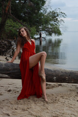 Young long-legged woman in a long red dress sitting on a wooden block on a sandy seashore