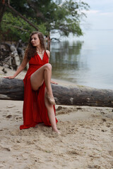 Young long-legged woman in a long red dress sitting on a wooden block on a sandy seashore