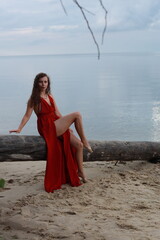 Young long-legged woman in a long red dress sitting on a wooden block on a sandy seashore