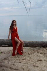 Young long-legged woman in a long red dress sitting on a wooden block on a sandy seashore