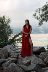 Pretty slim brunette woman in red dress standing full length on the rocks near green bushes