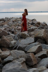 Beautiful slim young woman in long red dress standing on stones at the sea