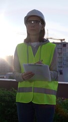 Woman an architect or engineer wearing safety hard hat and vest holding clipboard while inspecting...