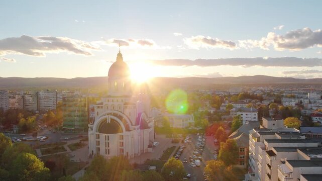 A cinematic aerial shot slowly orbits a majestic Orthodox cathedral in Bacau,Romania. The setting sun creates a spectacular lens flare and bathes the autumn cityscape in warm golden light.