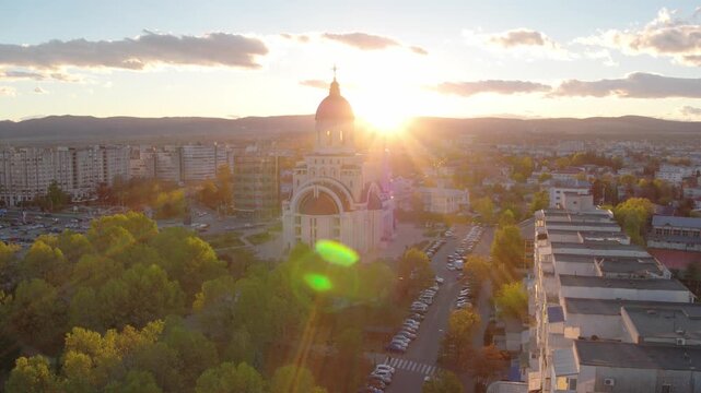 A slow-panning aerial shot of an Orthodox cathedral in Bacau, Romania. The setting sun creates a spectacular lens flare over the autumn park and cityscape.