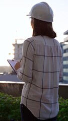 Woman construction engineer wearing white checked blouse and hard hat is making notes on a...