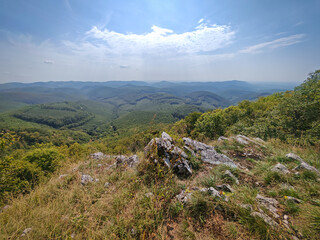 Panoramic view from the eastern cliff of Három-kő in the Bükk Mountains, Hungary. Rocky outcrop with vast green valleys and layered mountain ridges under a bright sky.