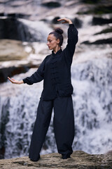 Fototapeta premium Woman practicing Tai Chi in nature, wearing a traditional black martial arts uniform, standing gracefully near a waterfall, focused on meditation and balance. Healthy lifestyle in asian culture