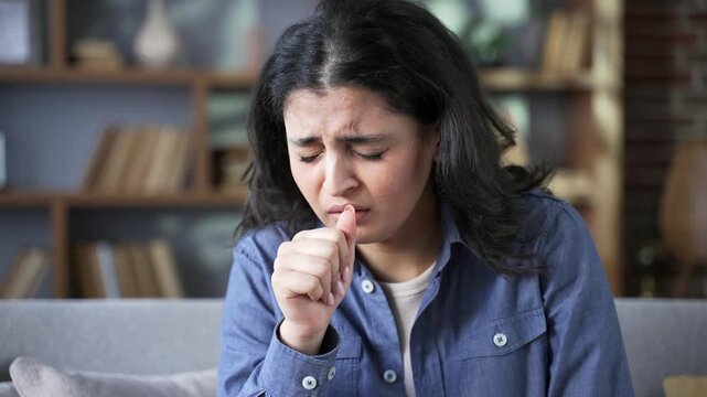 Sick young woman coughing while sitting on sofa in living room at home. Unhealthy adult female feels bad, holds her hand to her chest. The girl has cold, virus, flu, bronchitis or pneumonia. Close up
