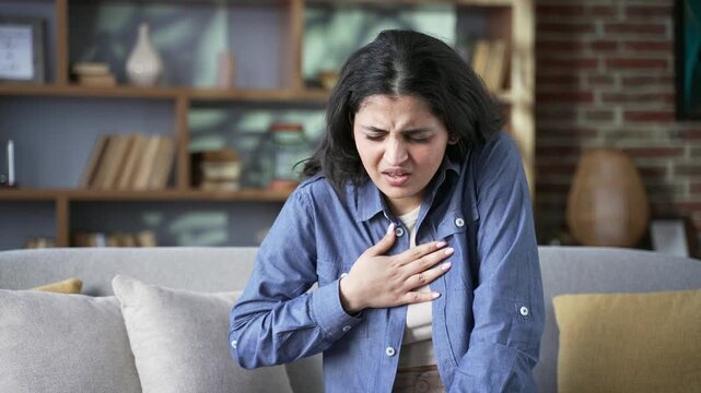 Young woman has a heart attack sitting on sofa in living room at home. Sick female suffers from tension in chest and massages the sore spot with her hand. She needs urgent medical attention. Close up