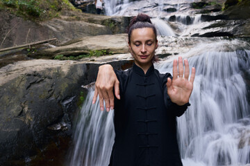 Woman practicing Tai Chi in nature, wearing a traditional black martial arts uniform, standing...