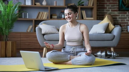 Confident female yoga trainer giving video call lesson using laptop sitting on mat on floor in living room at home. Young woman coach has online course engages in remote training or meditation session