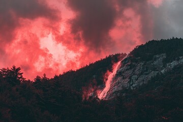 Red-hued clouds loom over a dark, forested mountain with a cascading waterfall