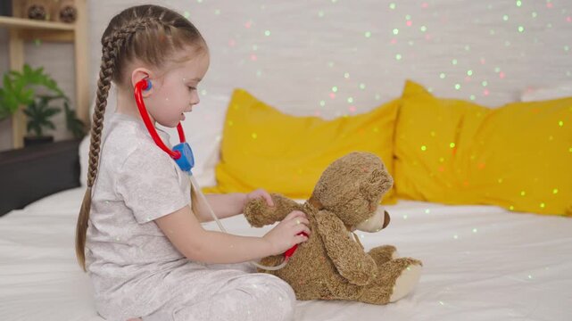 little child plays with teddy bear in patient in hospital, kid listens to stuffed toy with stethoscope, childhood dream becoming doctor, baby imagines himself as veterinarian, girl examining patient.