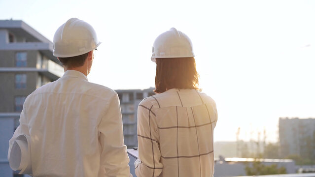 Two architects colleagues, wearing safety helmets and glasses, are examining blueprints at a construction site during sunrise or sunset, back view. Architecture and engineering