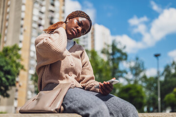 African American woman sitting in the city after work. She has pain in her neck and holding smartphone.