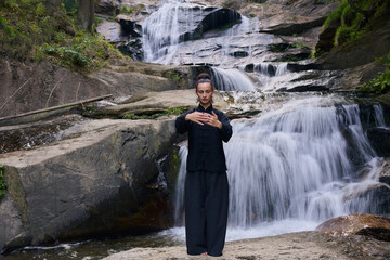Woman practicing Tai Chi in nature, wearing a traditional black martial arts uniform, standing...