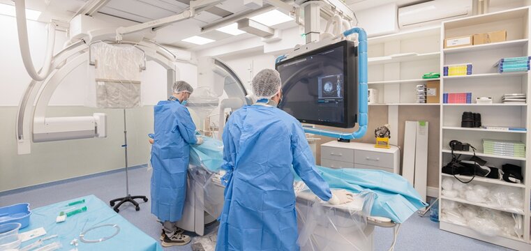 Two medical professionals wearing scrubs and hairnets meticulously examine brain scans displayed on a large monitor. They work in a bright, modern surgical suite at Saint Jude Hospital