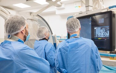 Three masked healthcare professionals in blue scrubs and hairnets intensely study a large medical scan. They are in a sterile operating room, likely performing interventional radiology