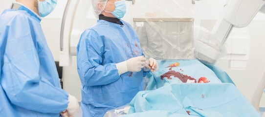 Medical personnel, wearing surgical scrubs and masks, work on a patient in an operating room. The table is covered with surgical instruments and bloody cloths during a medical procedure