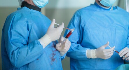 Two surgeons in a sterile environment, wearing masks and gloves, work with syringes filled with blood during a medical procedure, possibly in an operating room
