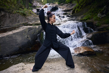 Obraz premium Woman practicing Tai Chi in nature, wearing a traditional black martial arts uniform, standing gracefully near a waterfall, focused on meditation and balance. Healthy lifestyle in asian culture