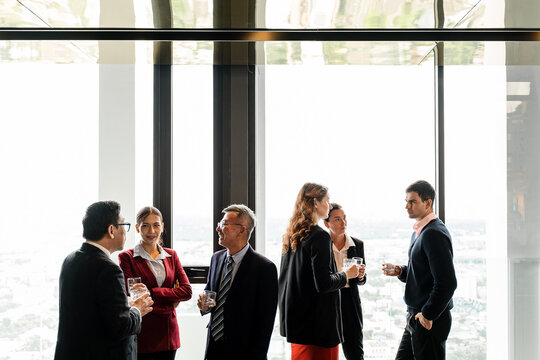 Business professionals networking in a modern high-rise with panoramic city views, chatting with drinks during a corporate event by large windows.